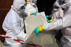 Three men in protective suits and gloves work on a large concrete slab during an asbestos removal process.